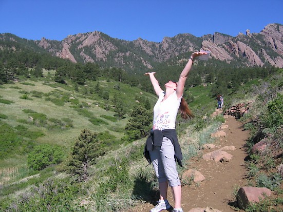 A white woman with arms to sky, standing on a trail leading up a mountain