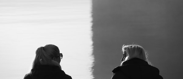 Two women sit side by side, one in white background and other in grey